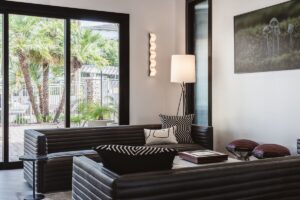 A view of the clubhouse at Borrego at Spectrum as redesigned by jeffrey allen design. A lounge area is next to a large sliding door that leads to the pool area with palm trees. There are two black leather sofas with a horizontal channeling, two wire stools with maroon padded upholstered seats, a small marble side table with rounded corners, and a coffee table with a designer Monopoly board. A zig-zag sconce and minimalist floor lamp provide additional lighting. Tall reeded glass windows let light in from the next space.